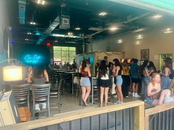 Indoor-outdoor urban café-bar with open garage door, neon wall sign, high-top tables, arcade games, industrial ceiling, and groups of people socializing