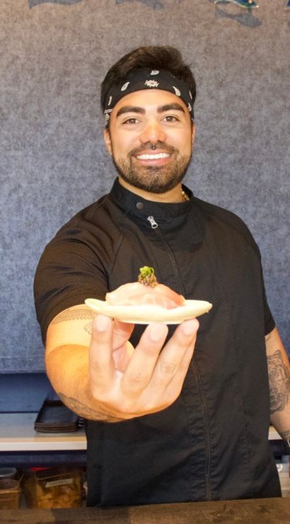 Smiling sushi chef in a black bandana presenting fresh nigiri on a small wooden plate at a sushi restaurant counter