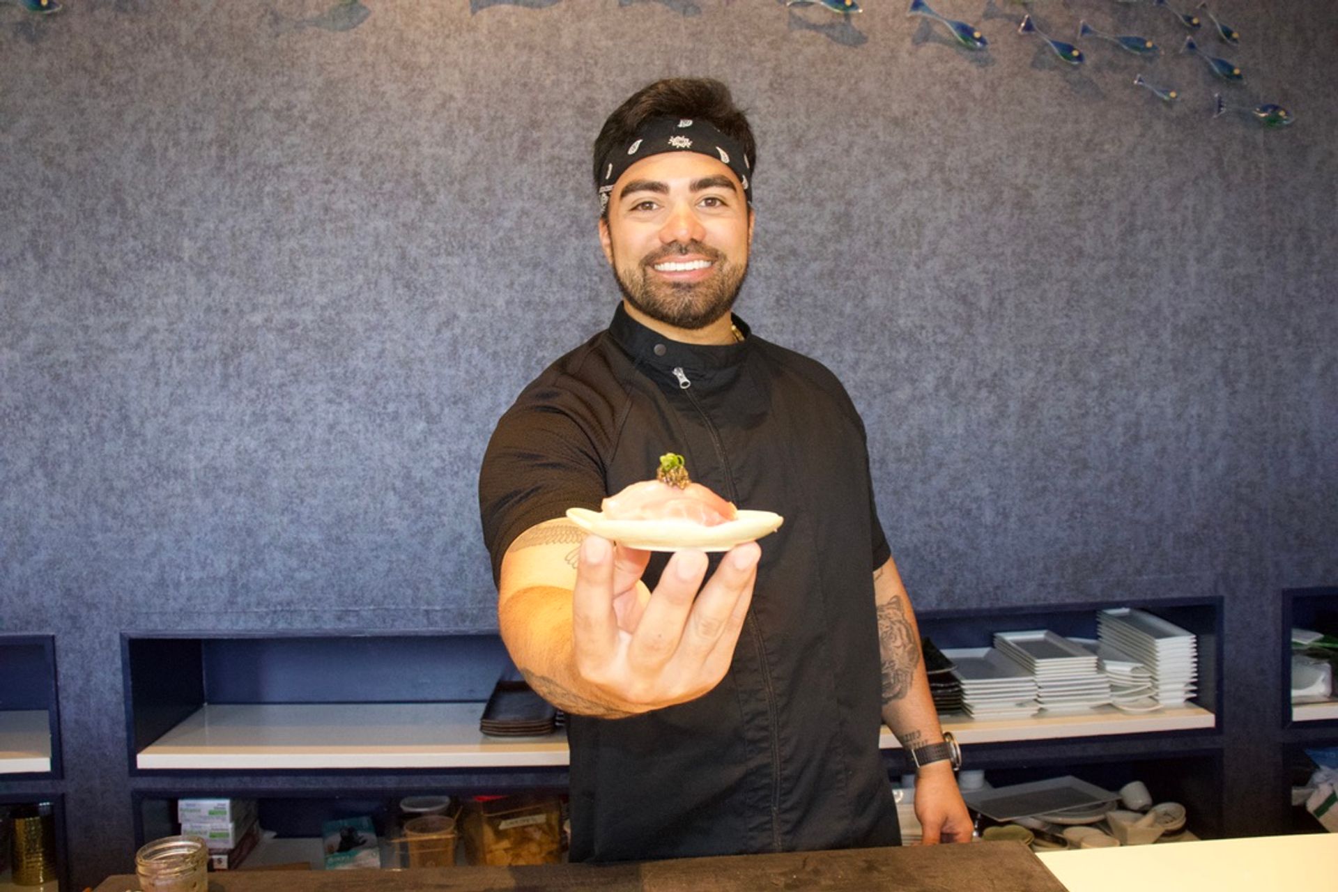 Smiling sushi chef in a black bandana presenting fresh nigiri on a small wooden plate at a sushi restaurant counter