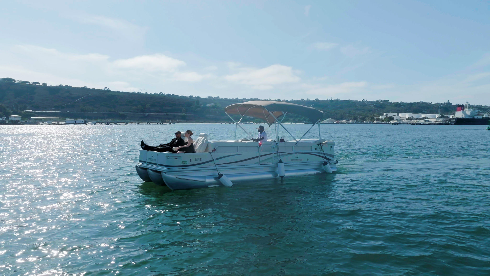 Leisure pontoon boat with passengers cruising a sunlit coastal harbor, rolling hills and port facilities in the background.