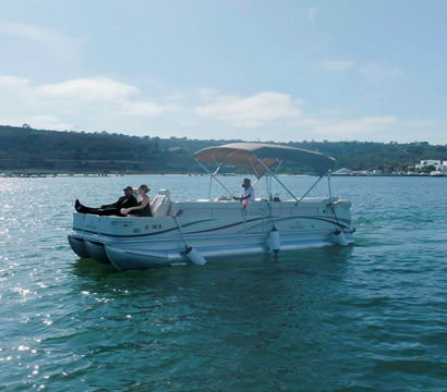 Leisure pontoon boat with passengers cruising a sunlit coastal harbor, rolling hills and port facilities in the background.