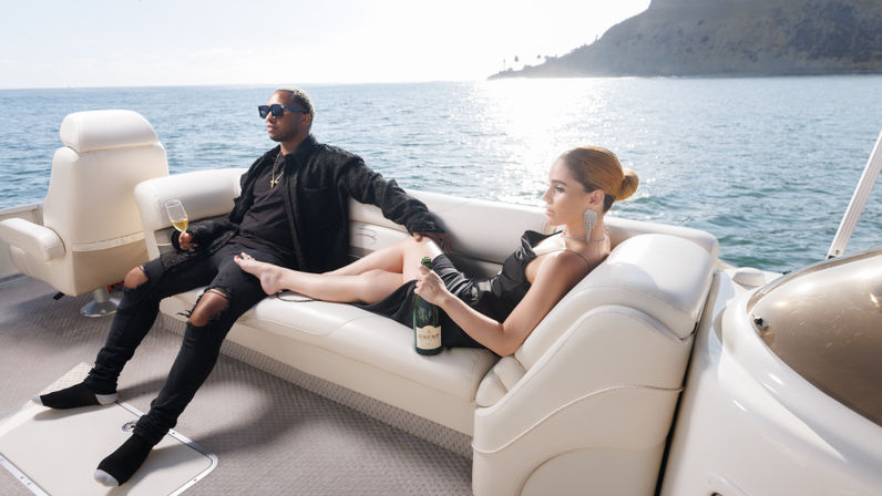Stylish couple lounging on a white yacht sofa, woman holding a champagne bottle and man with a flute, sunlit ocean and rocky coast in the background.
