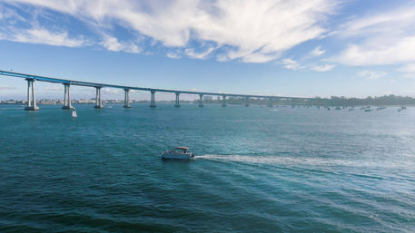 Motorboat cutting across turquoise bay leaving a white wake beneath a long curved coastal bridge, with a sailboat and distant skyline under wispy clouds.
