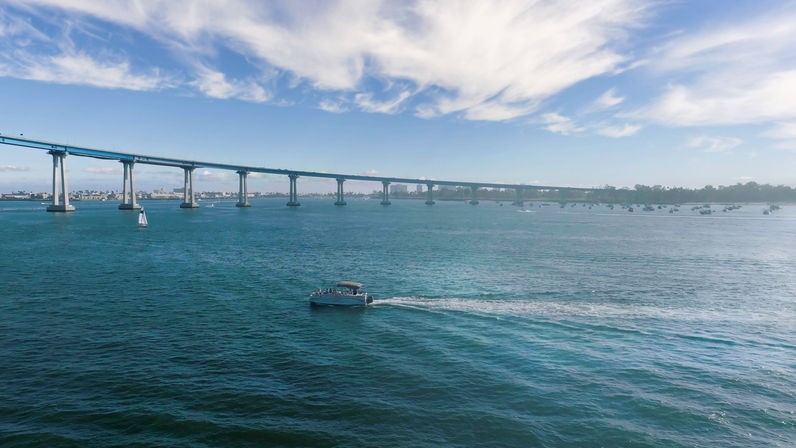 Motorboat cutting across turquoise bay leaving a white wake beneath a long curved coastal bridge, with a sailboat and distant skyline under wispy clouds.