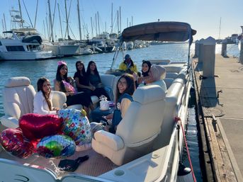 Smiling group of women enjoying a birthday with balloons and cake on a pontoon boat docked at a sunny marina with yachts and piers.