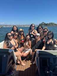 Boat party with a group of friends in swimsuits and sunglasses on a sunny day near a marina harbor, holding drinks and smiling under a clear blue sky