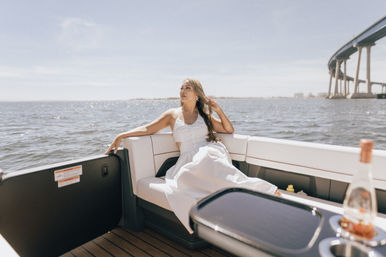 Woman in a white sundress lounging on a motorboat bench, sunlit bay waters with sparkling waves and a sweeping coastal bridge in the background under a clear blue sky