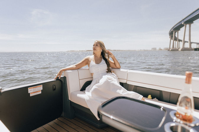 Woman in a white sundress lounging on a motorboat bench, sunlit bay waters with sparkling waves and a sweeping coastal bridge in the background under a clear blue sky