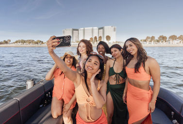 Six women in colorful swimsuits take a cheerful boat selfie with a coastal city skyline and tree-lined shoreline on a sunny day