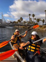 Two smiling people in yellow life jackets kayaking with bright orange paddles in a palm-lined marina, passing a pedestrian bridge and moored sailboats under blue sky and clouds.