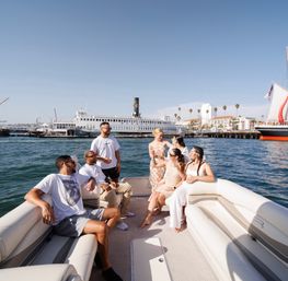 Friends relaxing on a pontoon boat in a sunny coastal harbor, toasting with drinks near a palm-lined waterfront and docked ships