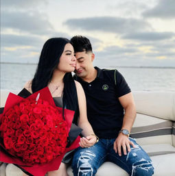 Romantic couple on a boat at sunset by the water, woman holding a huge bouquet of red roses while the man leans in, cloudy sky and calm sea in the background.