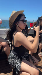 Person wearing a straw sun hat and sunglasses on a boat, kneeling in a black swimsuit with a patterned sarong, holding a phone with blue ocean and clear sky in the background and a red cup nearby.