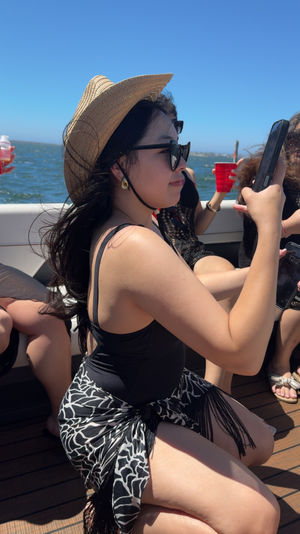 Person wearing a straw sun hat and sunglasses on a boat, kneeling in a black swimsuit with a patterned sarong, holding a phone with blue ocean and clear sky in the background and a red cup nearby.