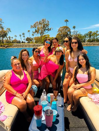 Smiling group of women in pink swimwear enjoying a sunny boat outing on a palm-lined coastal bay under a bright blue sky