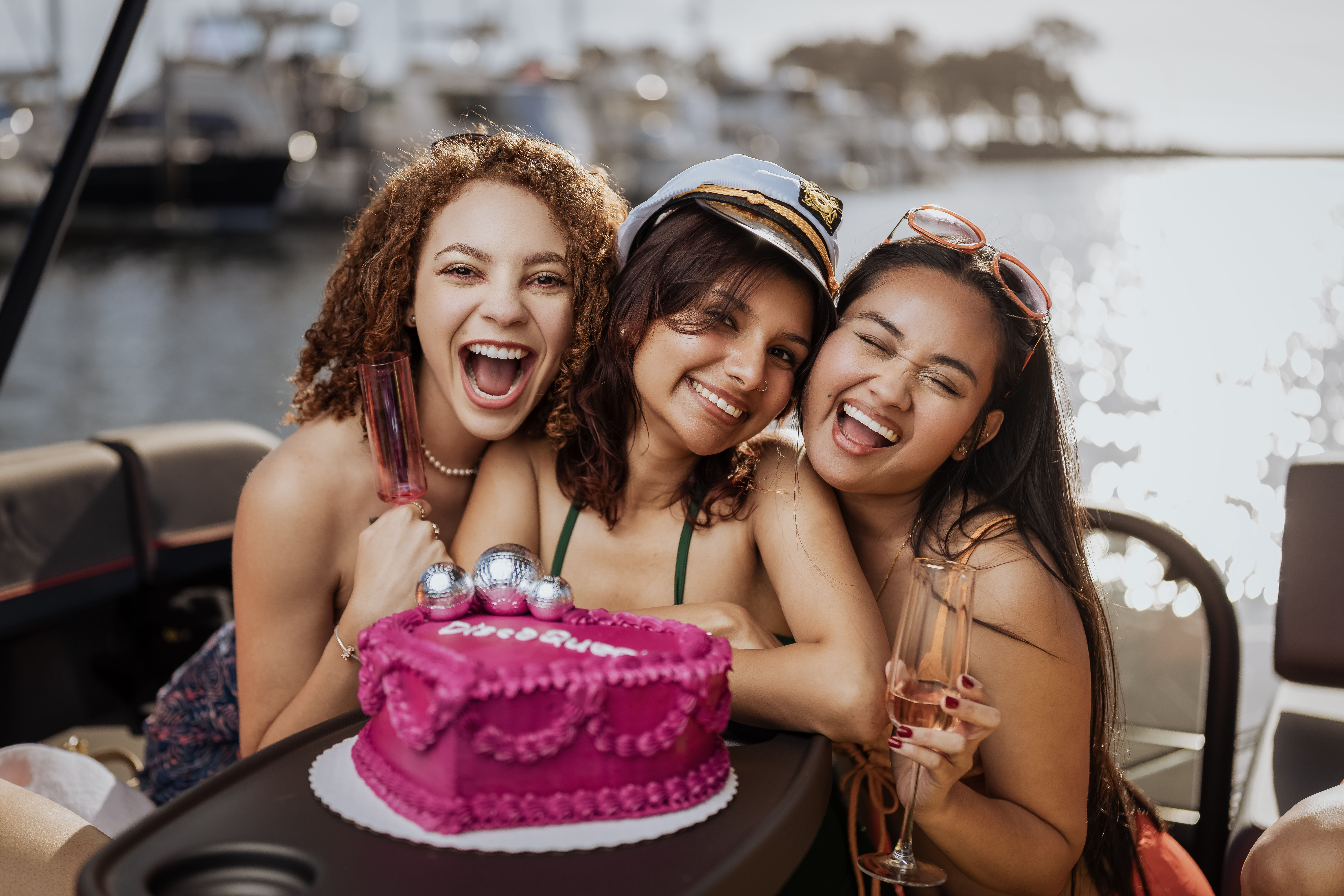 Three friends laughing on a sunlit boat at a marina, holding champagne flutes and leaning around a bright pink celebration cake topped with disco balls, one wearing a captain’s hat.