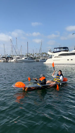 Two people paddling a transparent kayak with bright orange paddles and flotation buoys in a sunny coastal marina, yachts, sailboat masts and palm trees in the background.