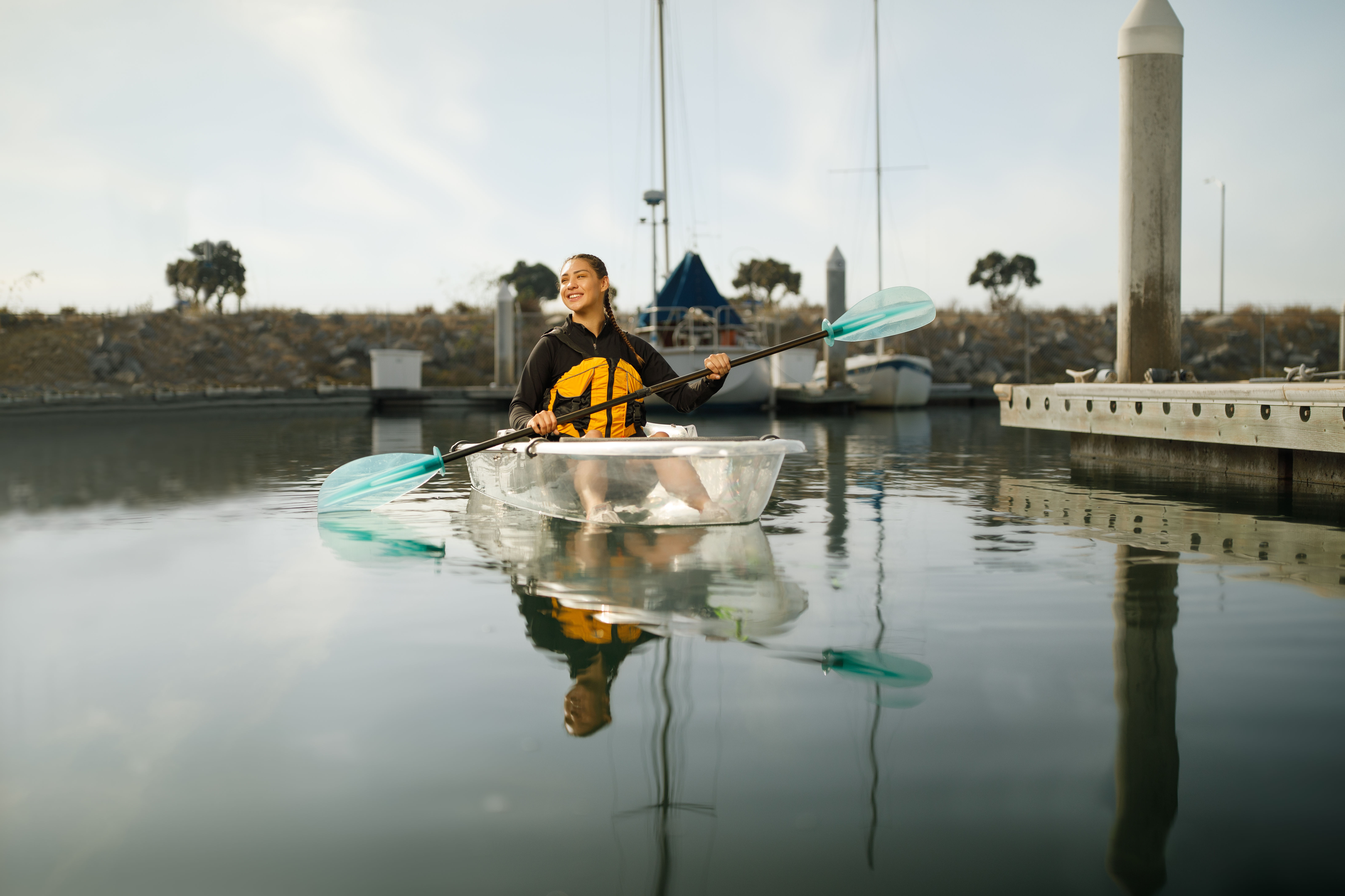 Smiling kayaker in a clear kayak wearing a yellow life jacket, paddling with turquoise blades in calm marina waters beside a wooden dock and moored sailboats.