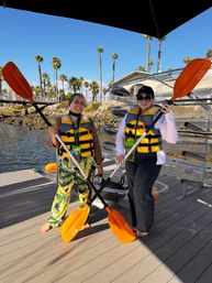 Two people in yellow life vests holding bright orange kayak paddles on a sunny coastal dock with clear kayaks and palm trees in the background