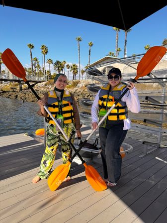 Two people in yellow life vests holding bright orange kayak paddles on a sunny coastal dock with clear kayaks and palm trees in the background