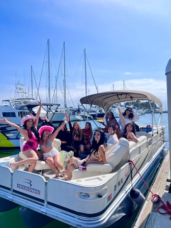 Group of friends enjoying a sunny pontoon boat party at a coastal marina — women in swimsuits and festive hats smiling on deck with sailboats and tall masts in the harbor background.