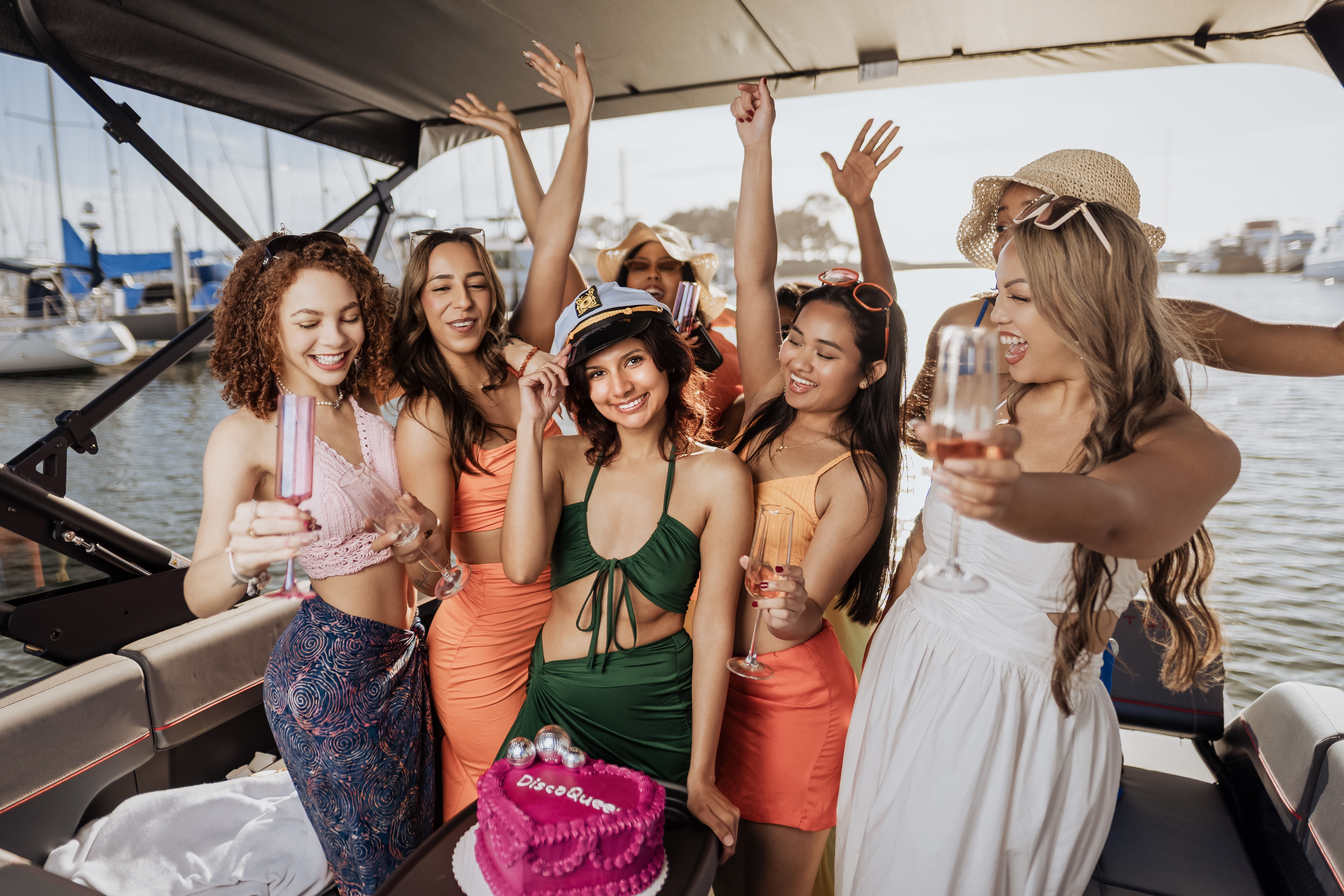 Women enjoying a sunny summer boat party at a marina, cheering with champagne around a pink cake while one wears a captain's hat.