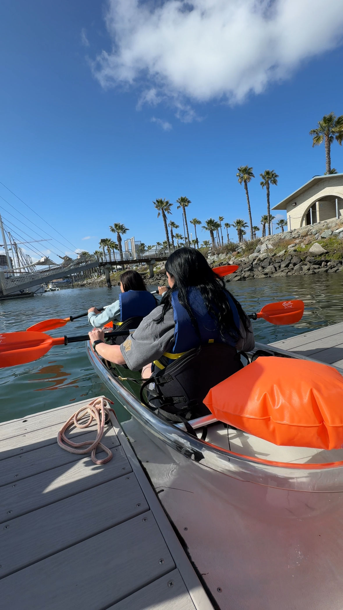Two kayakers in a clear tandem kayak with orange paddles and life jackets launching from a wooden dock at a sunny coastal marina lined with palm trees and a pedestrian bridge under a bright blue sky.