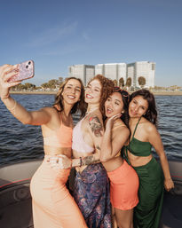 Four smiling friends in colorful summer outfits take a selfie on a boat in a sunny urban bay, with modern waterfront high-rise condos and shoreline trees behind them.