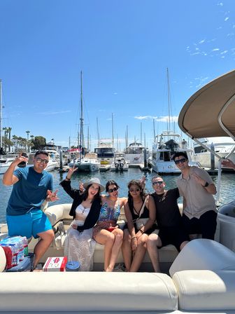 Six friends smiling and posing on a pontoon boat at a sunny coastal marina, sailboats and yachts docked behind them under a bright blue sky.