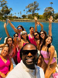 Cheerful boat selfie: man in sunglasses leads a lively group in pink swimsuits cheering on a sunny coastal bay with blue water and palm‑lined shore.