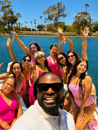 Cheerful boat selfie: man in sunglasses leads a lively group in pink swimsuits cheering on a sunny coastal bay with blue water and palm‑lined shore.