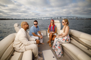 Four friends enjoying a pontoon boat ride on a coastal bay, chatting and laughing with a city skyline and arched bridge on the horizon under a dramatic cloudy sky.