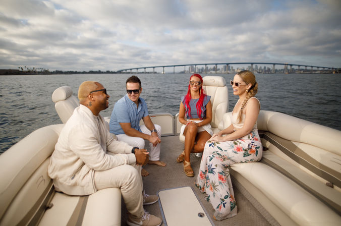 Four friends enjoying a pontoon boat ride on a coastal bay, chatting and laughing with a city skyline and arched bridge on the horizon under a dramatic cloudy sky.