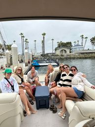 Seven friends laughing on a pontoon boat at a palm-lined coastal marina with a cooler on deck and an overcast sky
