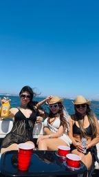 Three friends enjoying a sunny boat party at sea under a clear blue sky, wearing swimsuits, sunglasses and straw hats with red plastic cups, drinks on the table and a coastal city skyline on the horizon.