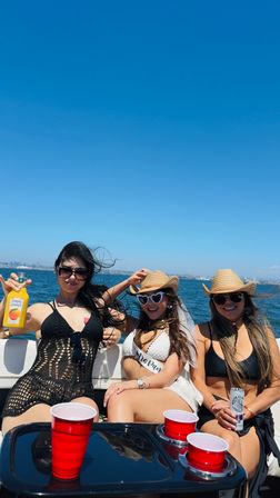 Three friends enjoying a sunny boat party at sea under a clear blue sky, wearing swimsuits, sunglasses and straw hats with red plastic cups, drinks on the table and a coastal city skyline on the horizon.