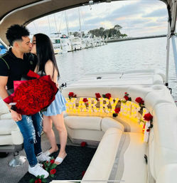Couple kissing on a yacht at a marina, woman holding a huge bouquet of red roses beside a lit "MARRY ME" sign and scattered roses on white cushioned seating, sailboats and harbor in the background.