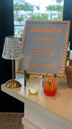 Tropical resort bar scene: wood‑beaded framed drinks menu listing Aperol Spritz and Hugo Spritz beside a small crystal lamp and two colorful cocktails on a white counter, with a pool and palm trees visible through the window.