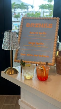 Tropical resort bar scene: wood‑beaded framed drinks menu listing Aperol Spritz and Hugo Spritz beside a small crystal lamp and two colorful cocktails on a white counter, with a pool and palm trees visible through the window.