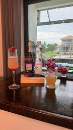 Festive cocktail display on a dark wood bar by a window overlooking a sunny waterfront canal with palm trees and dockside homes — two colorful drinks, bottles, orchids and a Happy 60th Birthday sign.