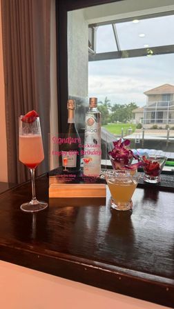 Festive cocktail display on a dark wood bar by a window overlooking a sunny waterfront canal with palm trees and dockside homes — two colorful drinks, bottles, orchids and a Happy 60th Birthday sign.