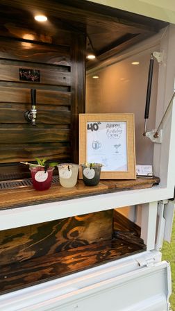Three colorful cocktails with garnishes on a wooden serving ledge of a mobile outdoor bar window, black draft tap and framed "40th Happy Birthday" menu in the background.