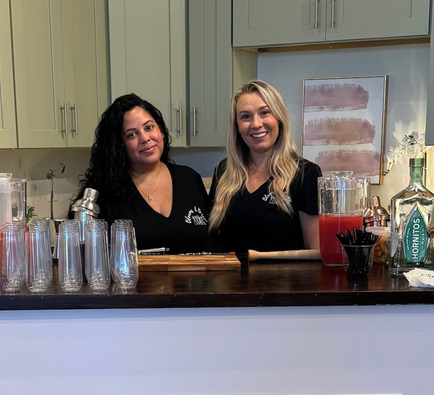 Two smiling bartenders behind a modern kitchen bar counter with stacked cocktail glasses, a shaker, wooden muddler, pitcher of red punch and a Hornitos tequila bottle — casual indoor cocktail setup for a small event.