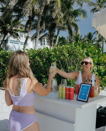 Two women clinking cocktails at a white beachside bar on a sunny tropical shoreline with palm trees, white sand, canned drinks and mixers on the counter.