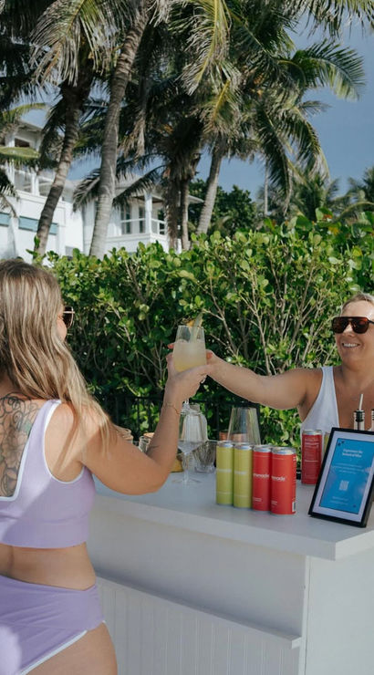 Two women clinking cocktails at a white beachside bar on a sunny tropical shoreline with palm trees, white sand, canned drinks and mixers on the counter.