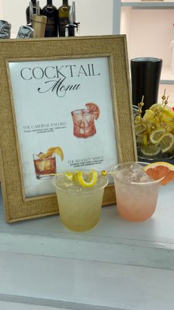 Framed cocktail menu on a white bar counter with two iced summer cocktails in plastic cups, garnished with a lemon twist and grapefruit wedge, jar of lemon slices and bar bottles in the background.