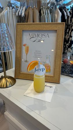Orange mimosa with an orange slice on a white counter in a boutique display, framed mimosa menu and neutral clothing racks in the background.