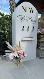 White VIP beverage tap wall with three chrome taps, pink and cream floral arrangements and pampas grass in a gold vase, glassware cart on a sunny outdoor tropical patio