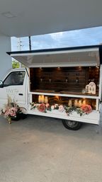 White mobile tap truck with wood-paneled serving counter, four taps, warm interior lighting and pink rose and greenery garland along the front, parked under a covered patio.