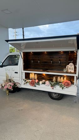 White mobile tap truck with wood-paneled serving counter, four taps, warm interior lighting and pink rose and greenery garland along the front, parked under a covered patio.